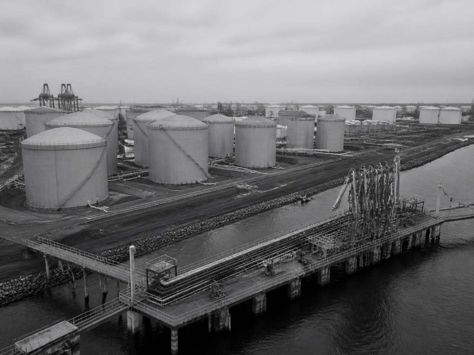 Monochrome photograph of a port tank terminal with storage tanks and a loading jetty