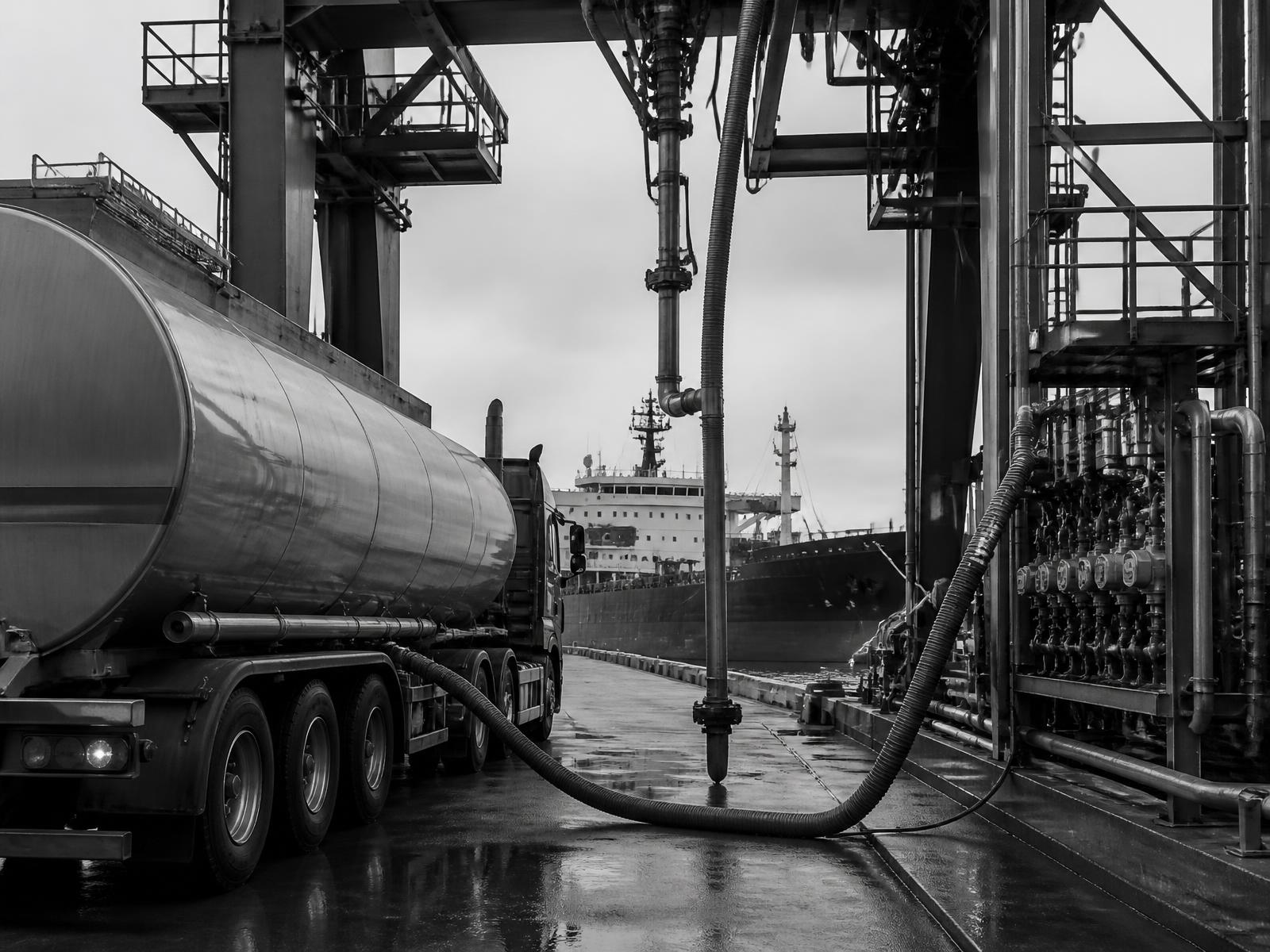 Monochrome photograph of a tanker truck loading at a terminal bay with a bulk vessel alongside